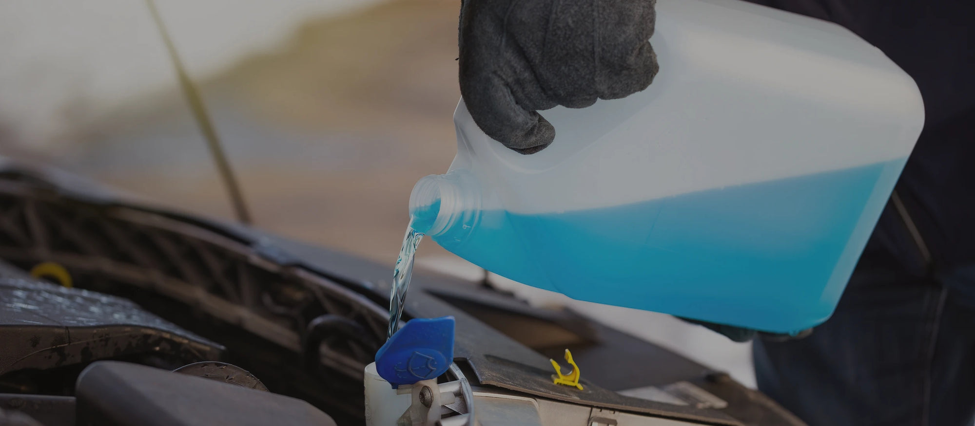 Person pouring blue liquid from a container into a car's windshield washer tank.